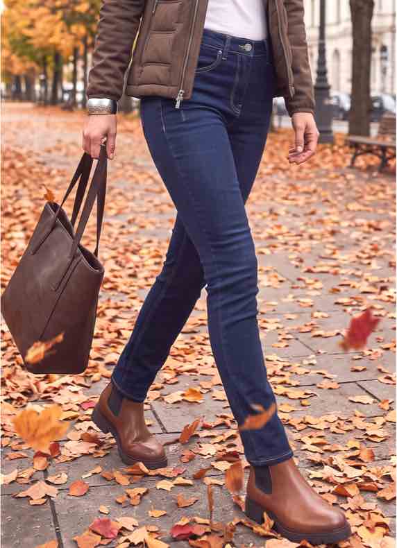 Frau in blauer Jeans und braunen Stiefeletten geht mit brauner Tasche über herbstliche Straße voller Laub